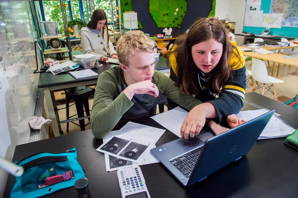 Shows two students working together at a desk with papers and a laptop