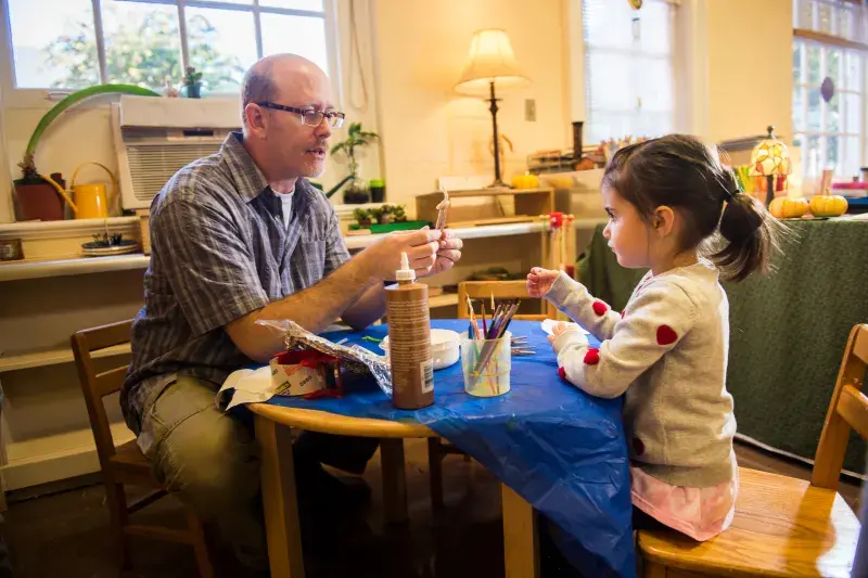Photo of a teacher sat with a young student helping with her art project.