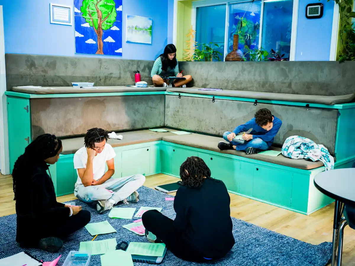 Photo of some older students sat on the floor by the bleachers with work spread out in front of them.