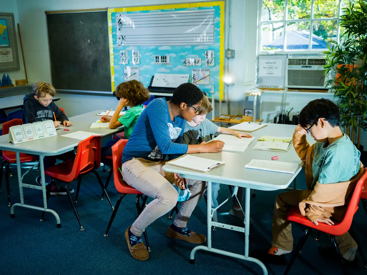 Photo of students in a classroom environment focused on their work. A teacher is sat assisting one of the students.