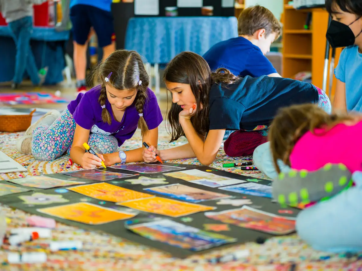 Children are sat on the floor with coloured markers, working on a project