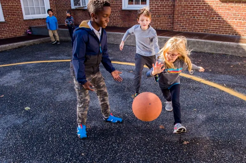Students are outside on a tarmac play area, playing a game with a ball (about the size of a football or volleyball).