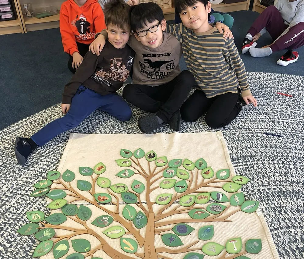 Three young students are posing for the camera in front of a large crafted tree with illustrations on the leaves.