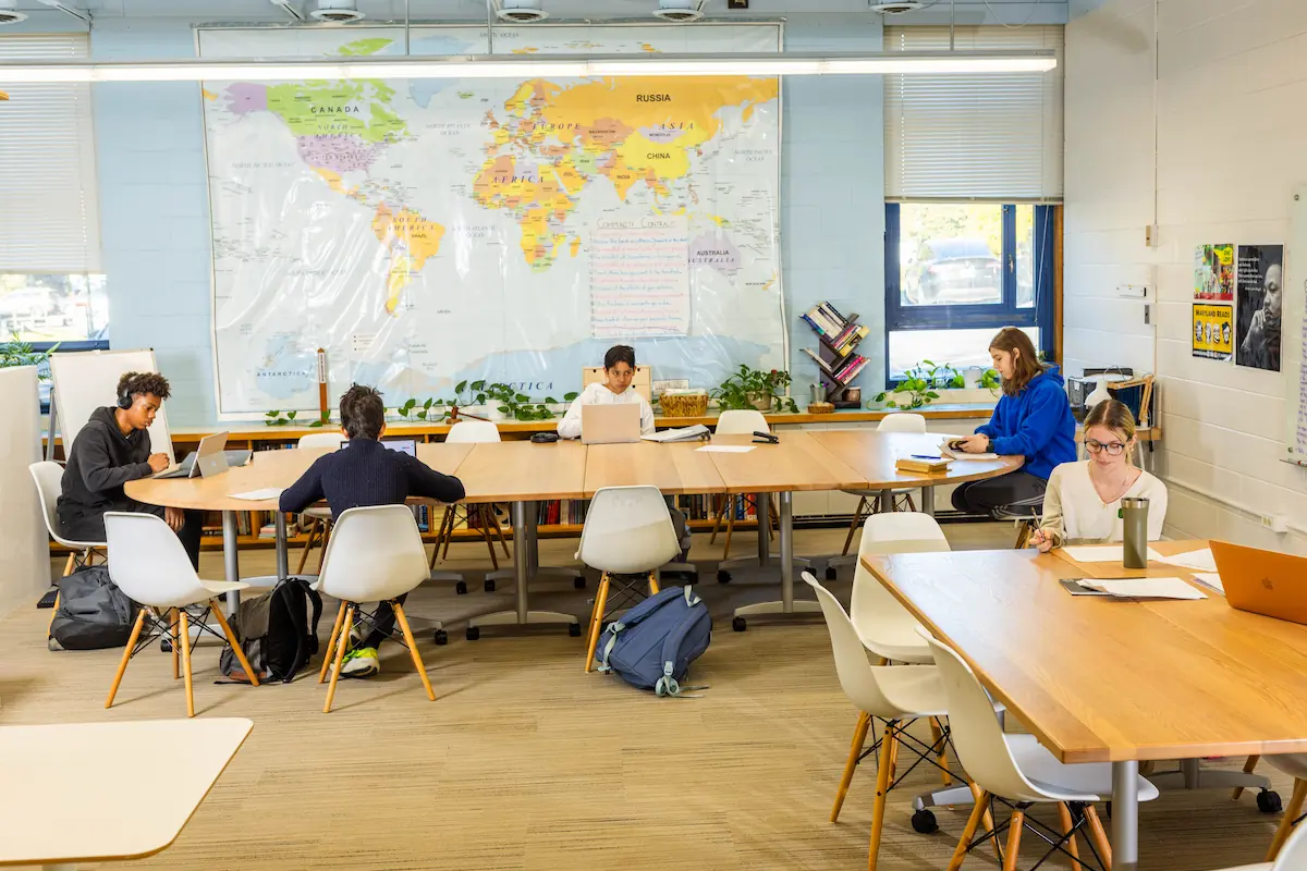 A group of students are sat at a large communal table in front of a world map.