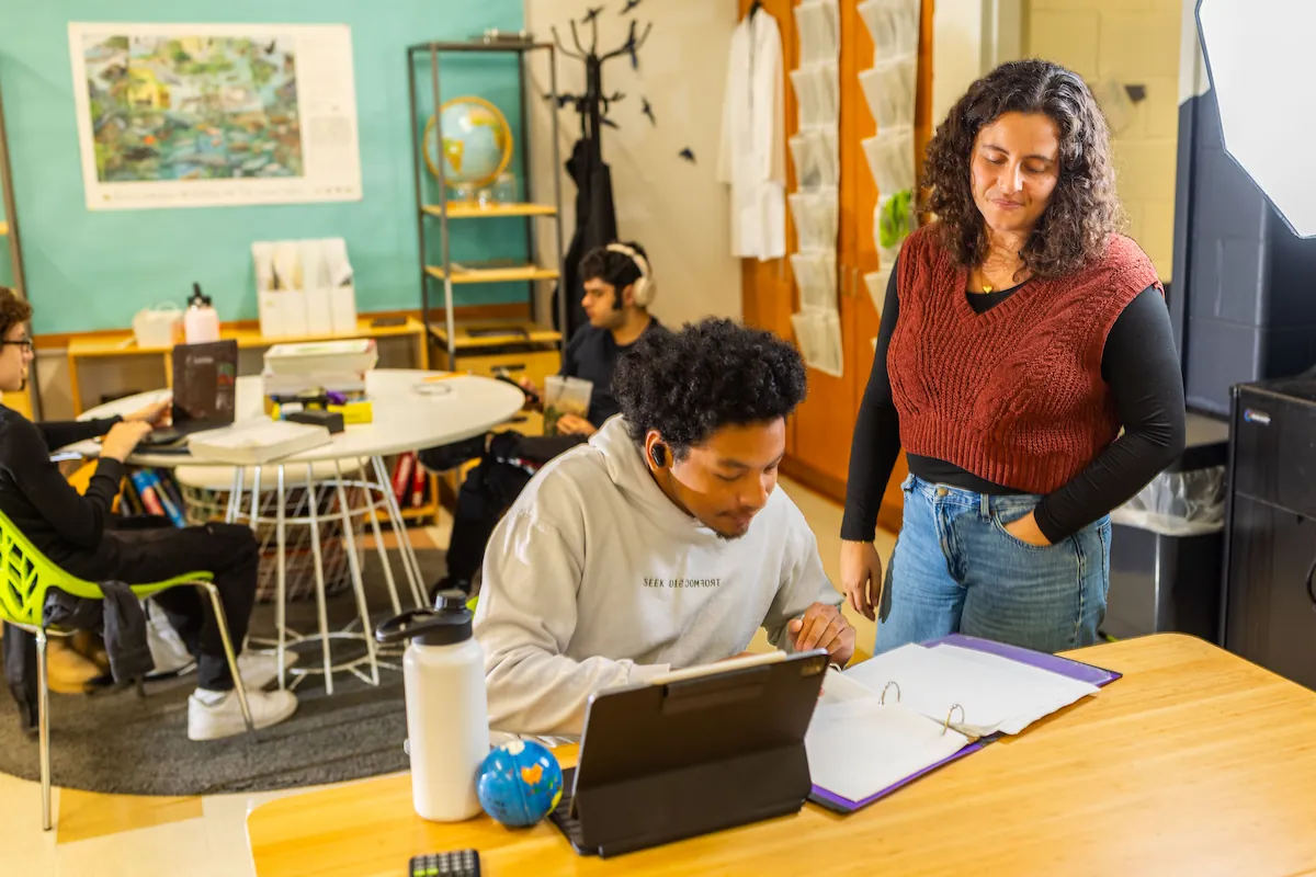 A senior student is working intently with headphones on while a teacher looks over his shoulder.