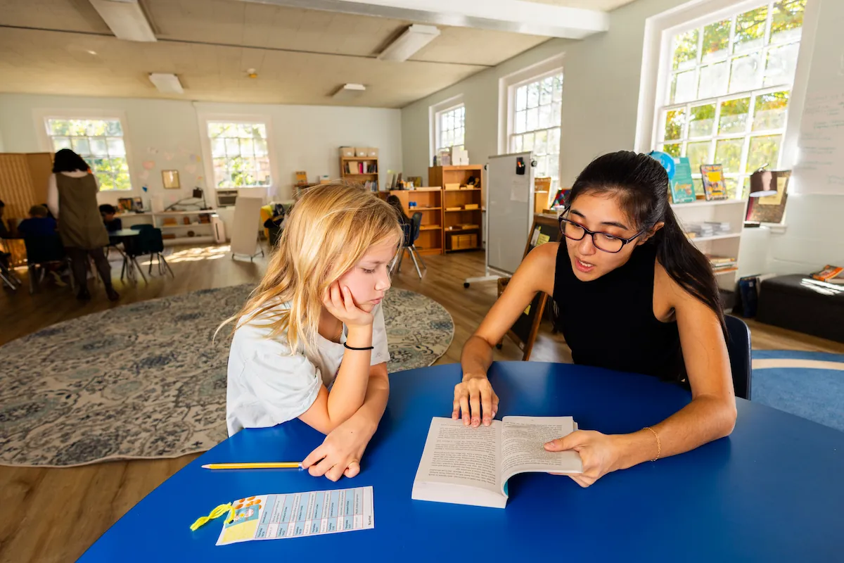 A student sits at a table with a teacher reading from a book