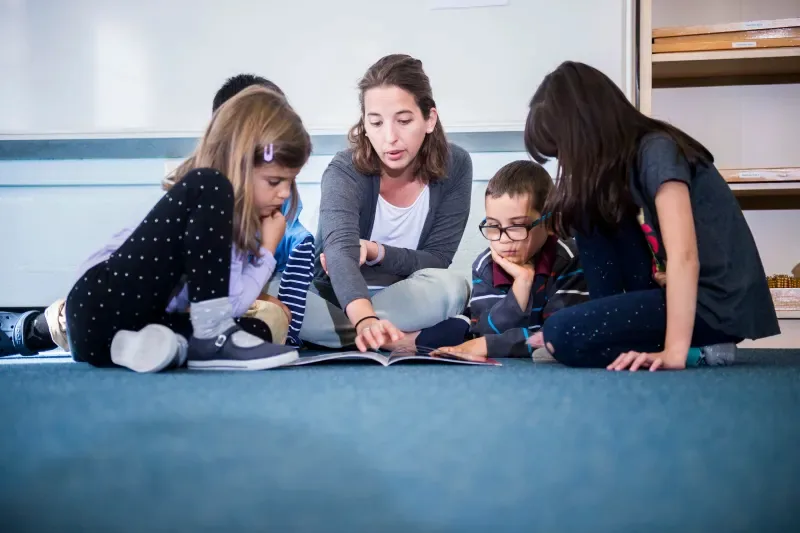 A teacher is sat on the floor with a group of students looking at a book.