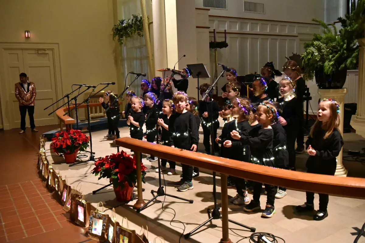 Photo of a group of young children singing on stage wearing decorative head pieces.