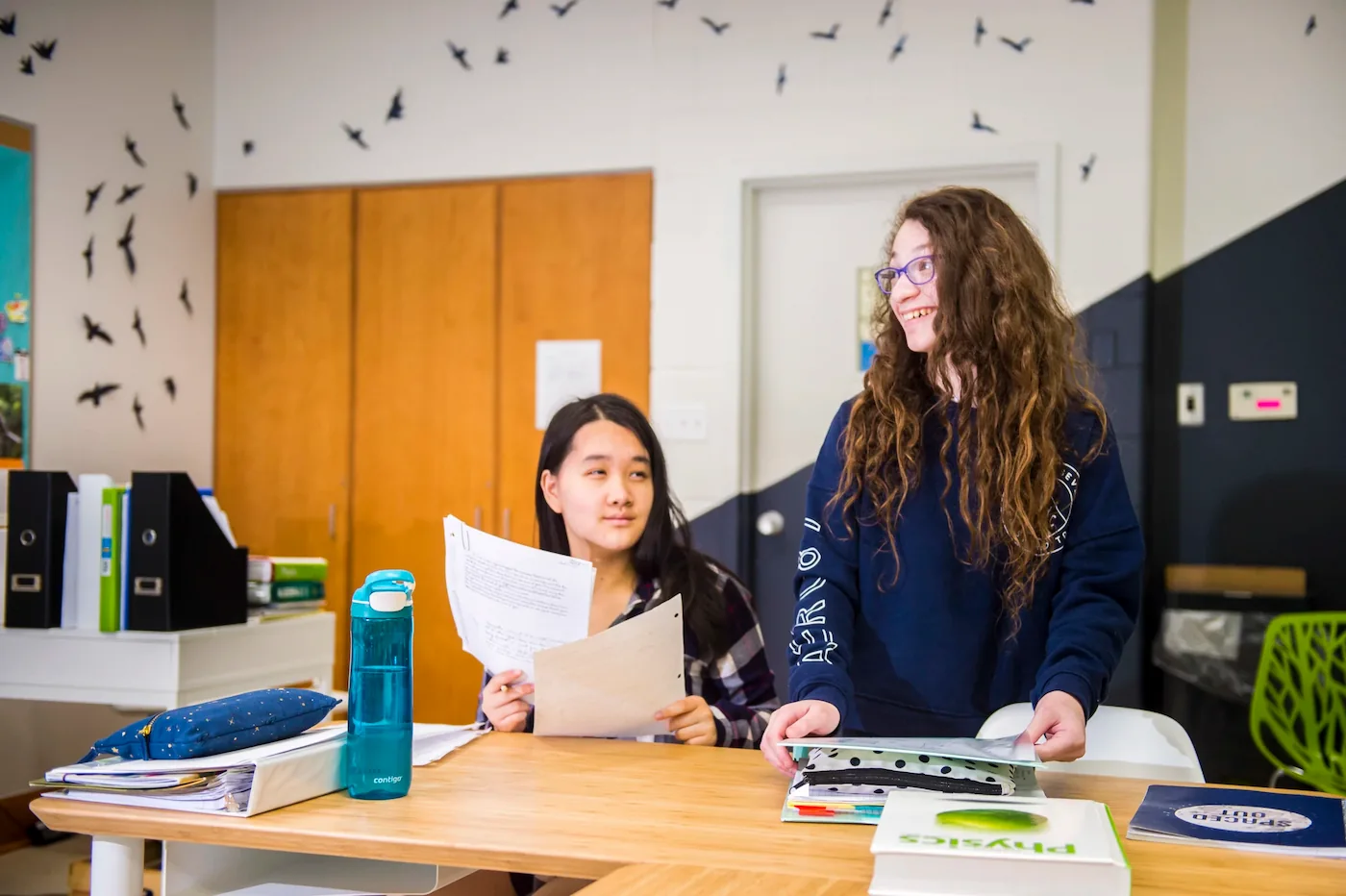 One student is holding papers from a research project, and another is smiling and in discussion with someone off screen.