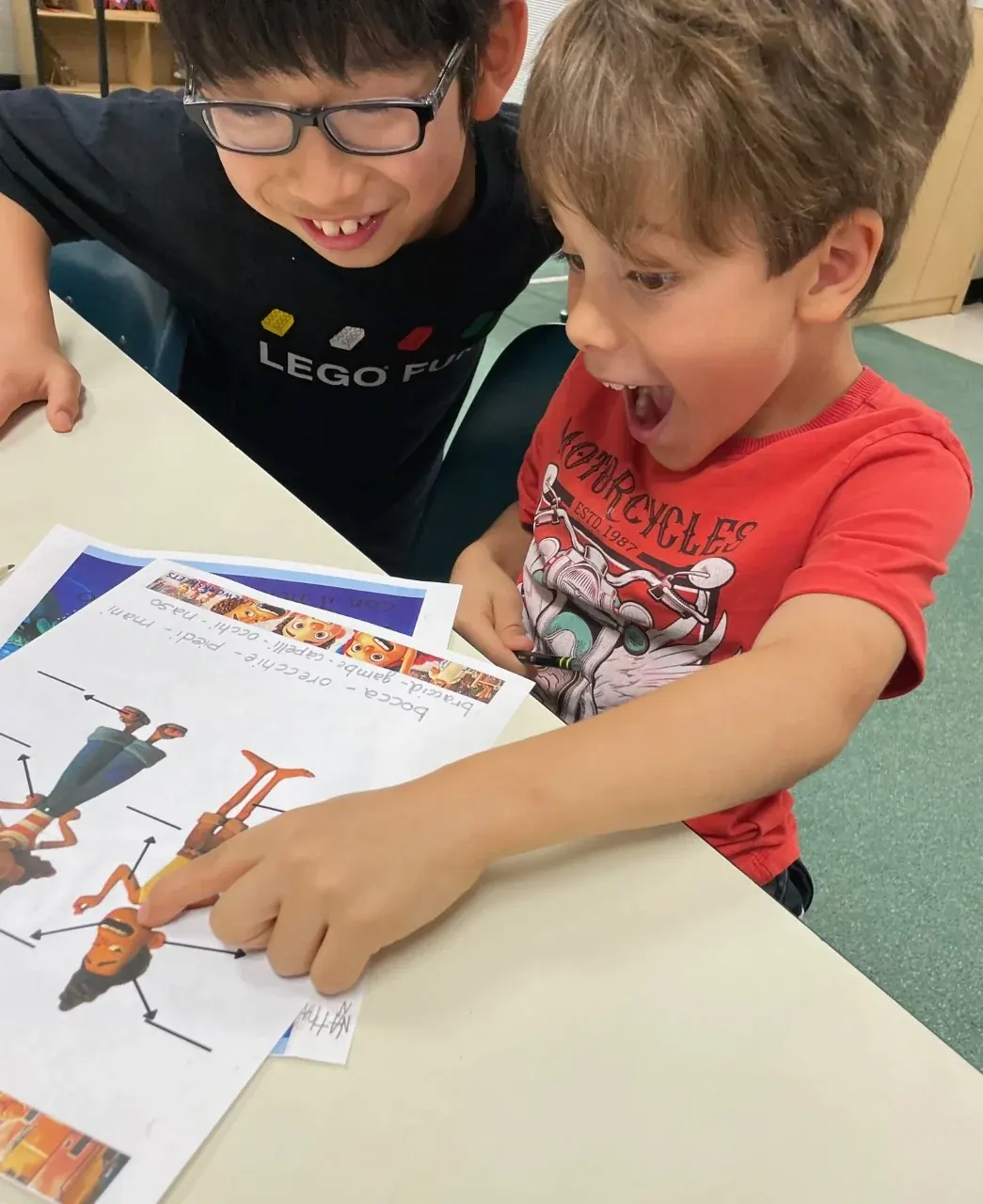 Two enthusiastic young students are sat together, one pointing to the head on an illustrated figure in his workbook