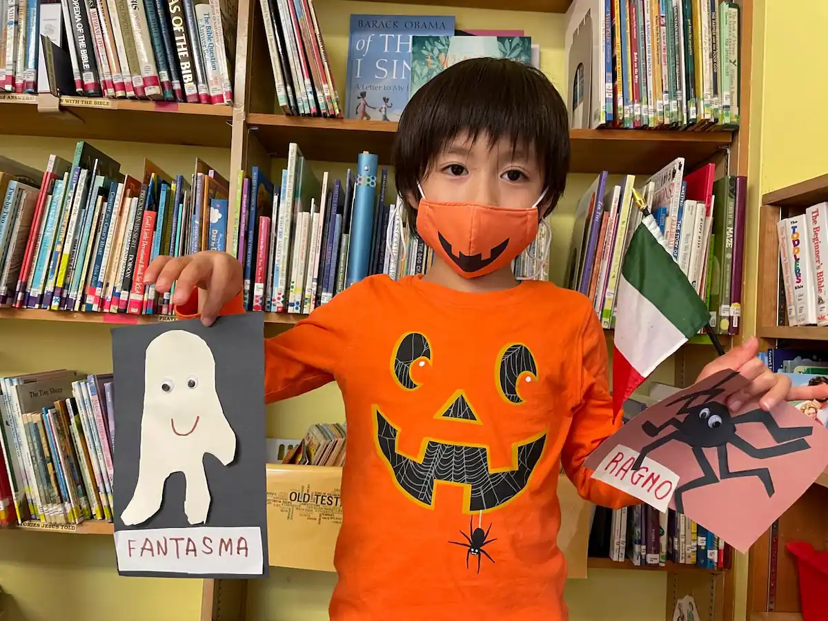 A student in a halloween shirt and mask holds up their spooky artwork.