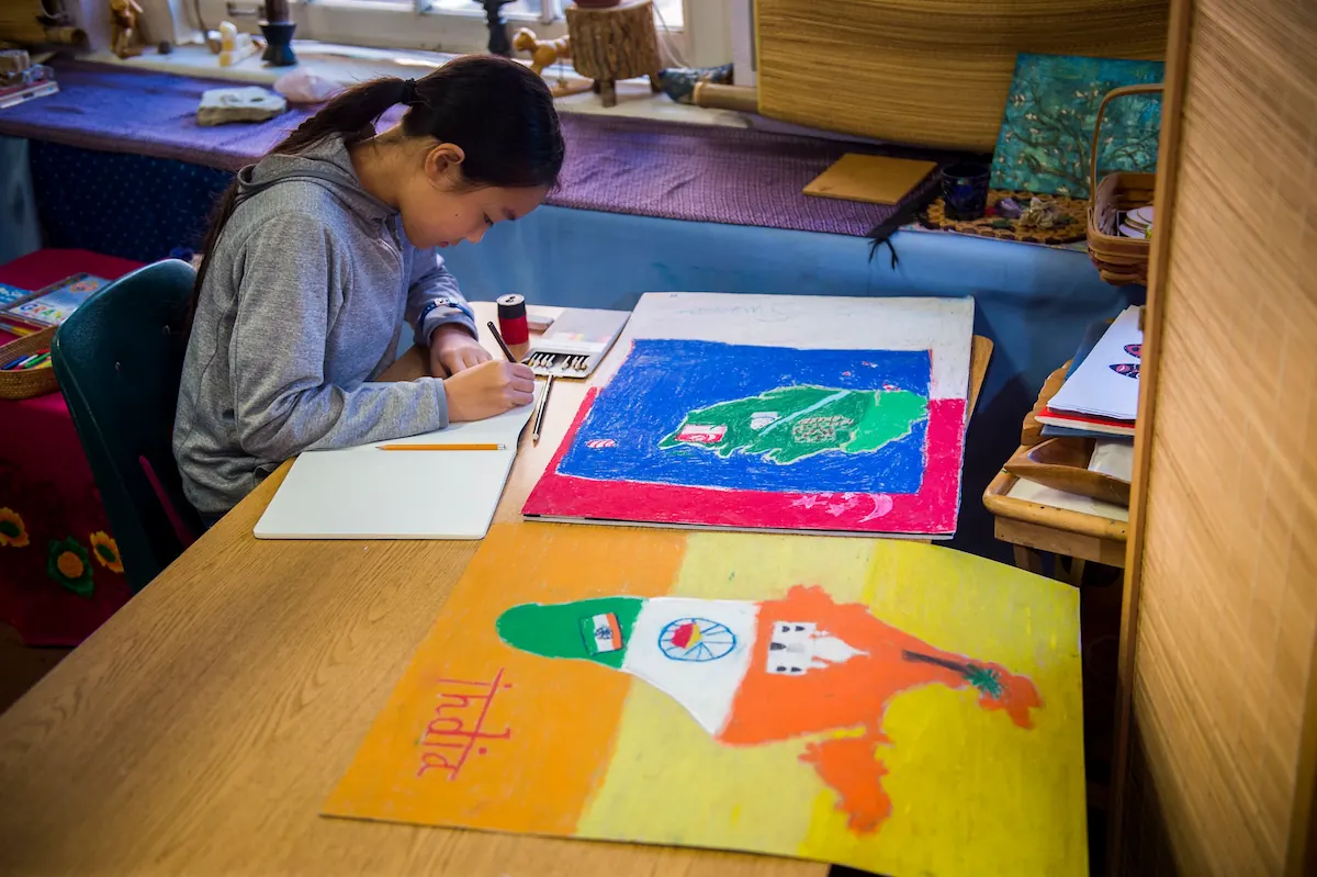 A student is using coloured pencils on a project. Her artwork is spread out across the desk.