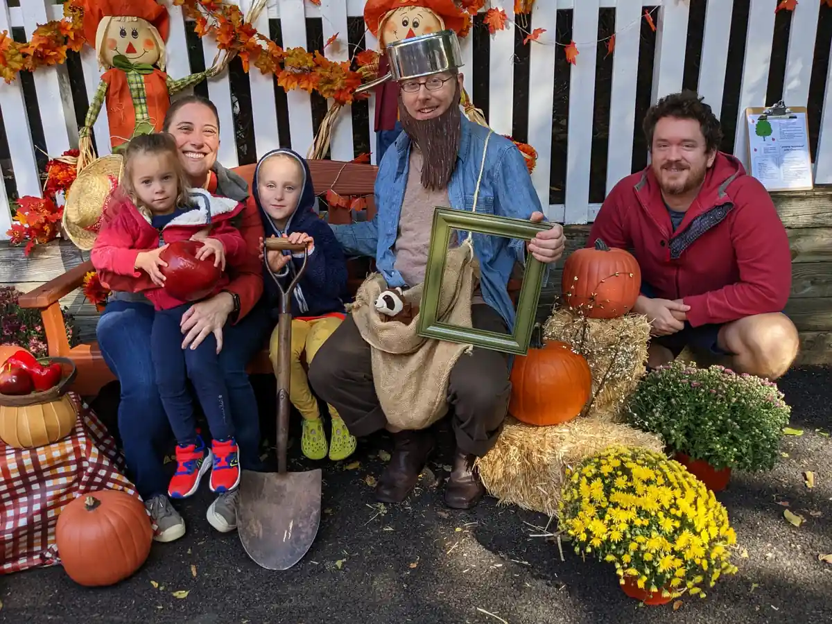 An adult in a silly outfit is posing with a family. The scene has pumpkins, flowers and bales of hay for decoration.