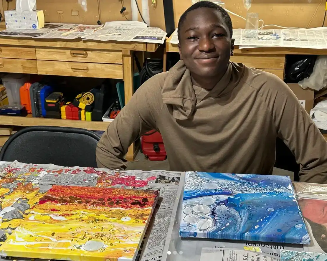 A smiling student is sat in the studio in front of two colourful painting featuring strong texture.