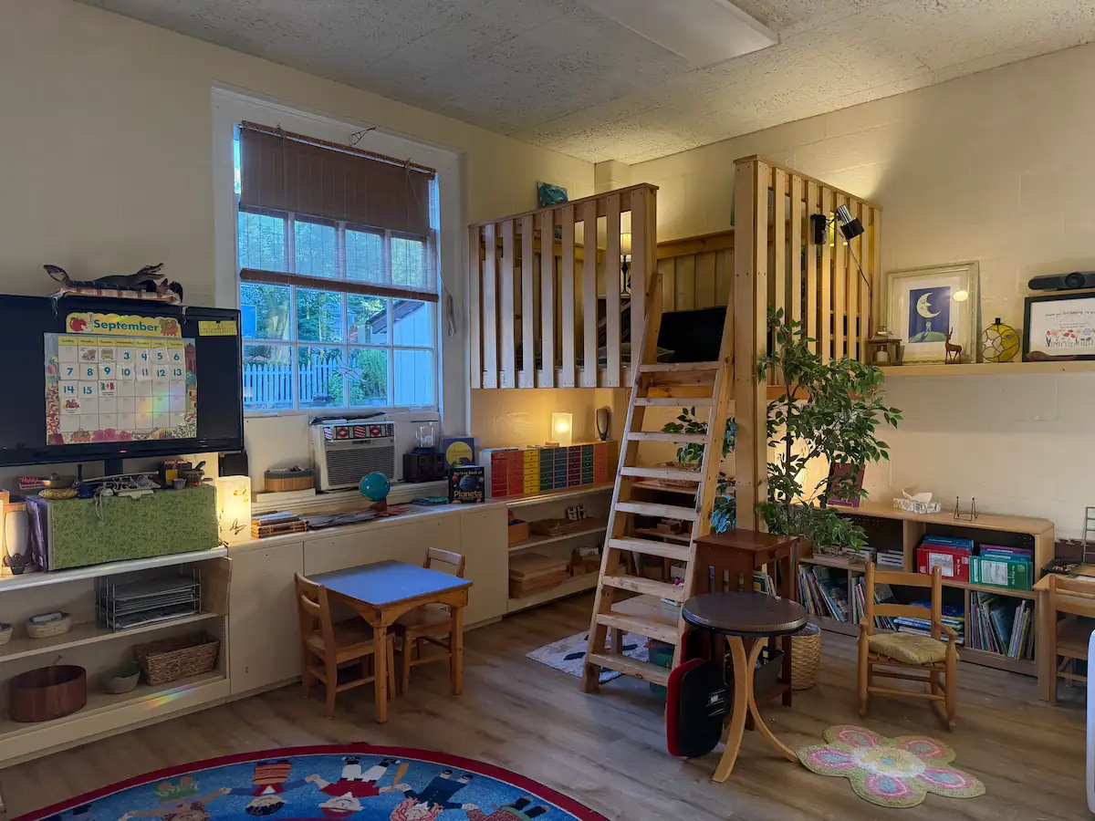 Montessori classroom corner with reading loft, ladder, low shelves, plants, and child-sized tables.