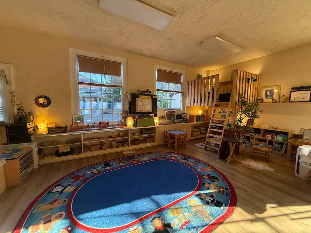 Wide view of a Montessori room with low shelving, blue oval rug, and wooden loft.