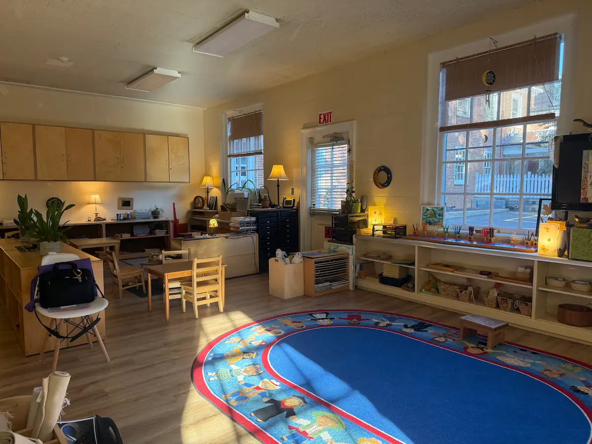 Sunlit Montessori classroom with cabinets, open shelves of materials, and a large rug.
