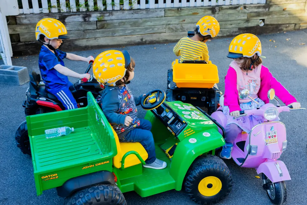 Four children are playing in toy vehicles wearing yellow helmets.