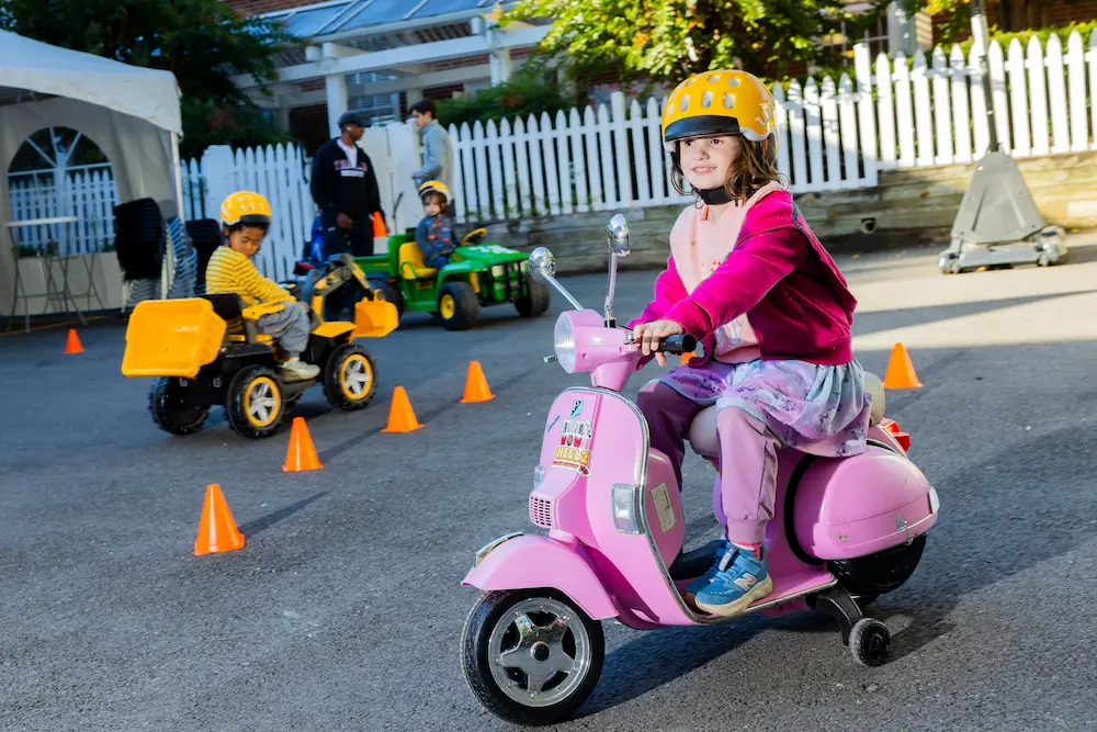 A young girl is riding a toy pink moped around a playground.